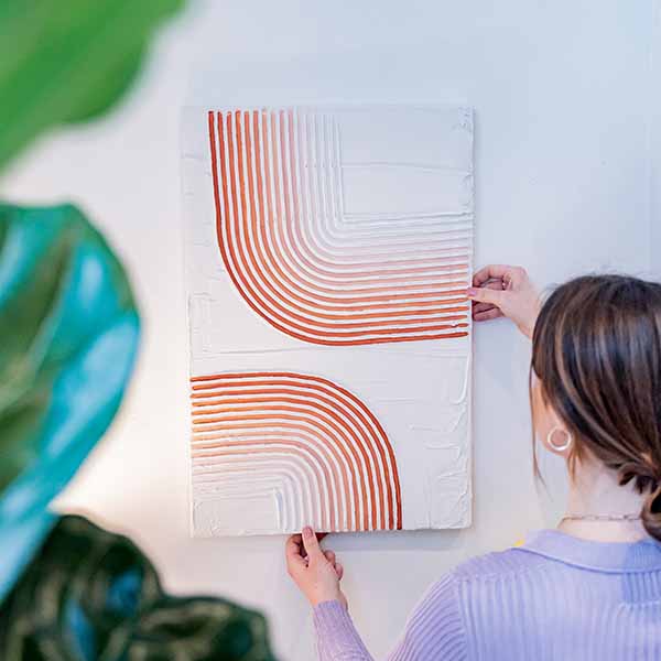 A woman hangs a textured, abstract artwork with curved orange lines on a white wall made of Sculpd Textured Canvas Kit