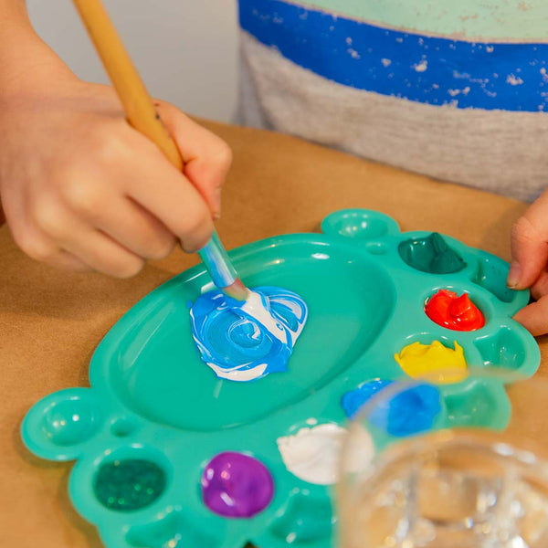 A child's hand swirling paint with a wooden brush on a Sculpd Kids Painting Kit Paint Palette