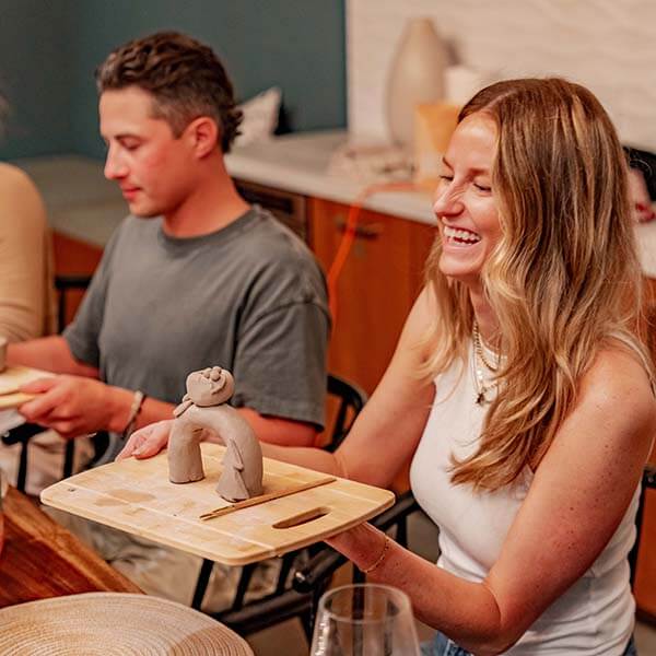 A woman laughs while holding a wooden board with a clay sculpture, enjoying a Sculpd Pottery Kit night