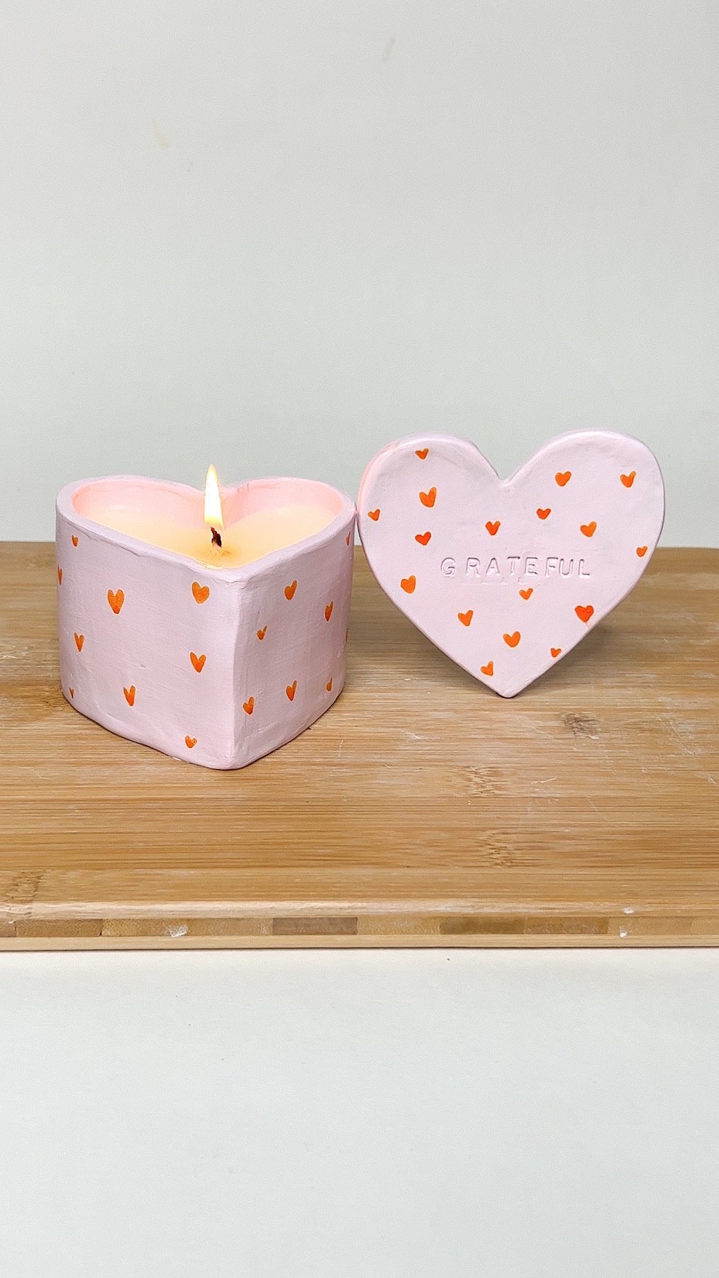A finished and lit Sculpd Candle Making Kit Candle vessel shaped like a heart with a heart-shaped cover on the side stamped with the word Grateful